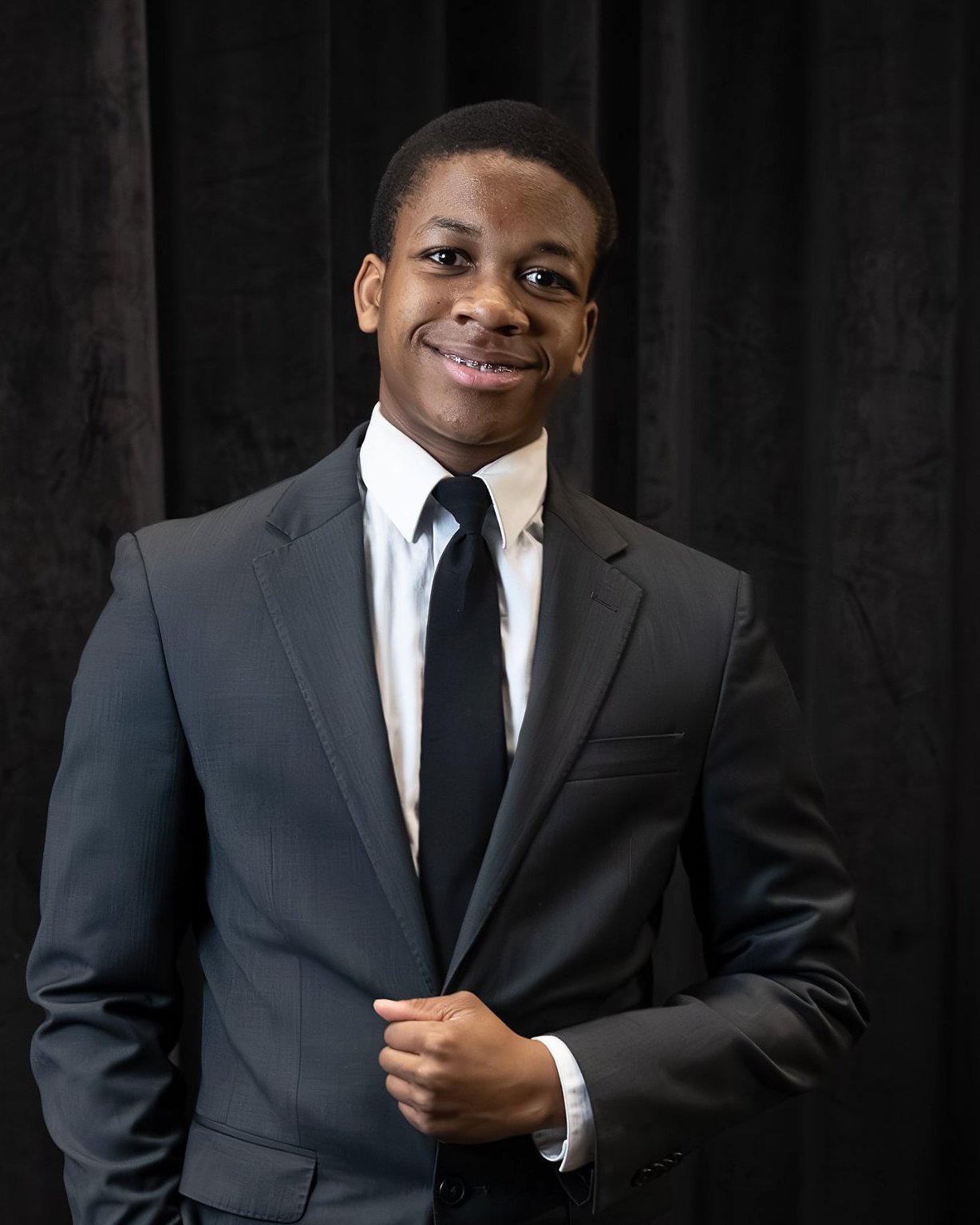 Young man in a dark suit and tie smiling in front of a black curtain